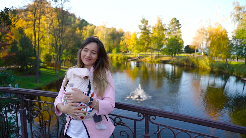 A young mother stands on a bridge on the background of a beautiful lake with her baby in a sling, she smiles and robs her baby.