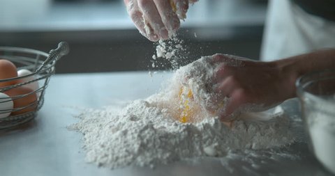 Pastry chef mixes a raw egg with flour on a metal counter top next to a metal basket of eggs and a bowl of flour, in soft light, in slow motion. Closeup shot in 4K on Phantom Flex