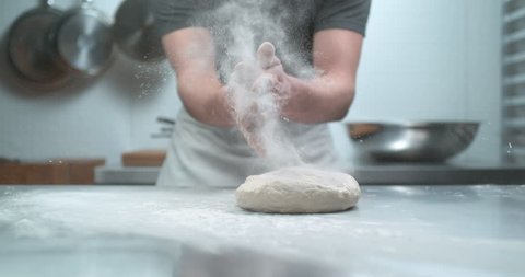 Pastry chef in white apron claps hands as flour bursts off into a cloud over a metal kitchen work counter with dough and flour. Shot 4K Phantom Flex.