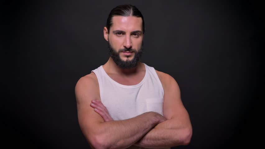 Serious strong bearded caucasian man is standing with crossed arms isolated on black background, wearing white shirt, chilling
