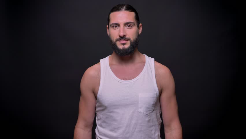 Cute strong fit caucasian guy is demonstrating like sign while standing in dark studio wearing white shirt and showing his muscular hands indoor