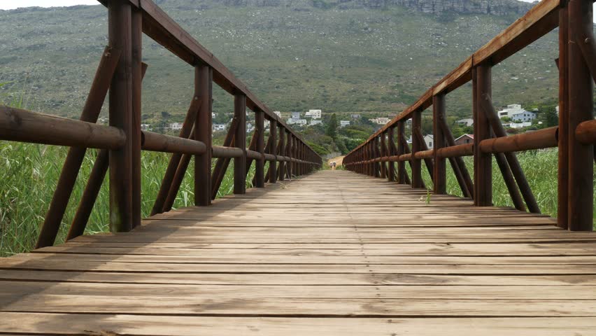 Wooden walkway bridge across wetland long grass conservation area, close to ground shot of wooden boards rising to bridge perspective with two people walking & distant houses against mountain