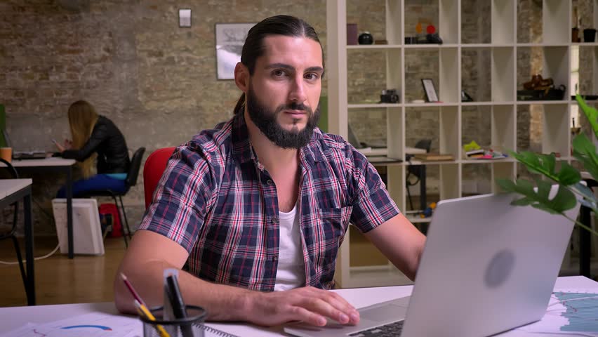 Smiling caucasian man with nice beard and long hair is relaxing during working time and looking at camera with smile while sitting at his workplace in modern office isolated