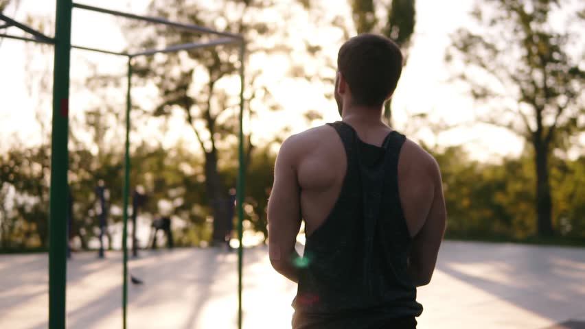 Fit man in black shirt doing pull-ups on horizon bar on sports ground with trees and sun shines on the background