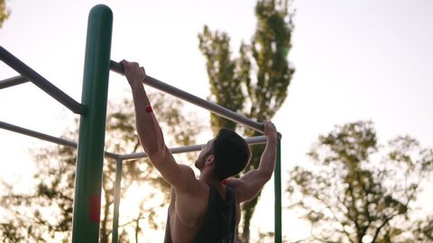 Fit man in black shirt doing pull-ups on horizon bar on sports ground with trees and sun shines on the background