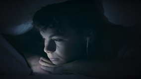 Night shot of teen boy with headphones using tablet pc under blanket. Happy child playing with cellphone or smartphone on a bed. Light reflection on face cute young boy - close up. 