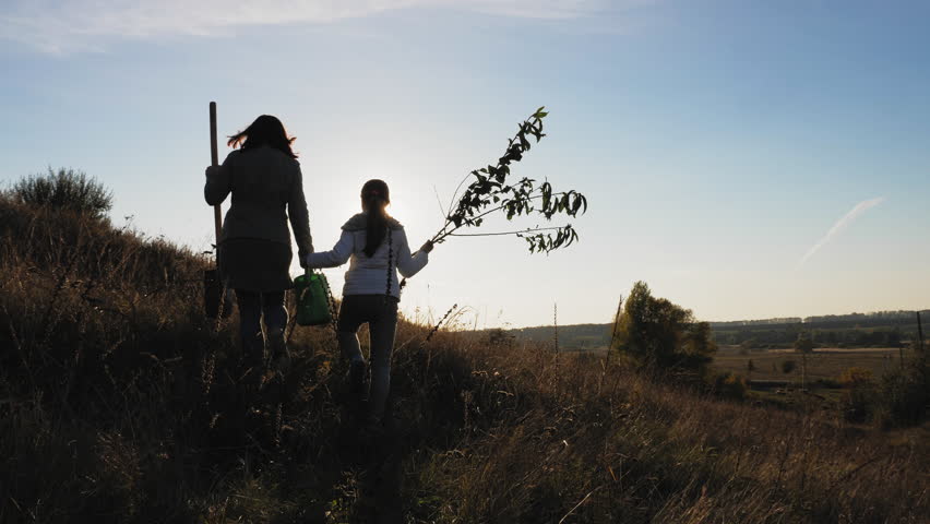 Rear view: Mom and daughter are going going to plant a tree. Carry a seedling, shovel and watering can. Silhouettes in a beautiful hill on a sunset background