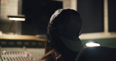 Young Asian woman sound producer bobs her head to music she is working on at night in audio studio over professional mixing boards. Medium shot on 4K RED camera.