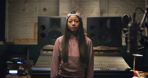 Portrait of urban girl with long hair and backwards baseball cap in pink hoodie and jeans in front of professional mixing board in a recording studio. Medium shot on 4K RED camera.