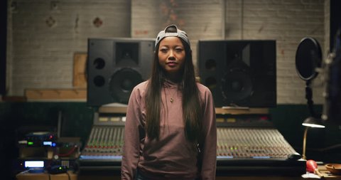 Portrait of young millennial Asian woman smiling with long hair and backwards baseball cap in pink hoodie and jeans in front of professional mixing boards in studio. Medium shot on 4K RED camera.