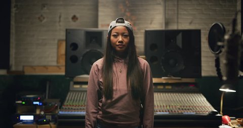 Portrait of young millennial Asian woman with long hair and backwards baseball cap in pink hoodie and jeans in front of professional mixing boards in studio. Medium shot on 4K RED camera.