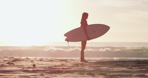 Young, beautiful, blonde surfer woman stands with her surfboard on the beach with the sea and surf in the background in dawn light, in Australia. Medium shot in 4K on a RED camera