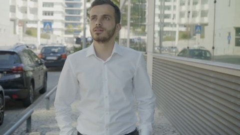 Caucasian young man walking along street in thought. Manager in white shirt looking up and down and contemplating on project. Tracking camera, slow motion. Weighing pros and cons concept 