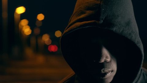 Hooded black man alone in the dark raising head looking at camera- city night