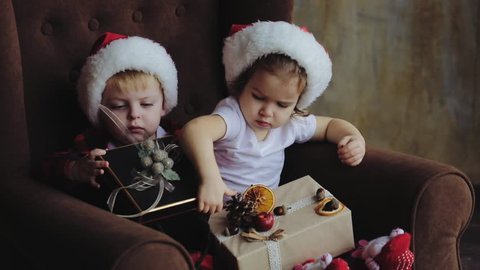 Young children, brother and sister, sit in the living room under the Christmas tree in Santa Claus hats and open presents. The traditional decoration of the room with light bulbs. Family concept