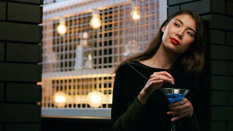 Attractive Asian woman with a cocktail in hand looks around the corner waiting for someone in interior cool bar with soft interior lighting. Medium shot on 4k RED camera.