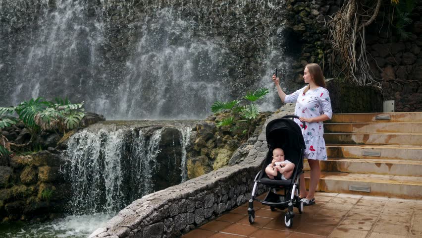 A young mother walking with her baby near the waterfall takes pictures on her smartphone for social networking and posting online while traveling with her family.
