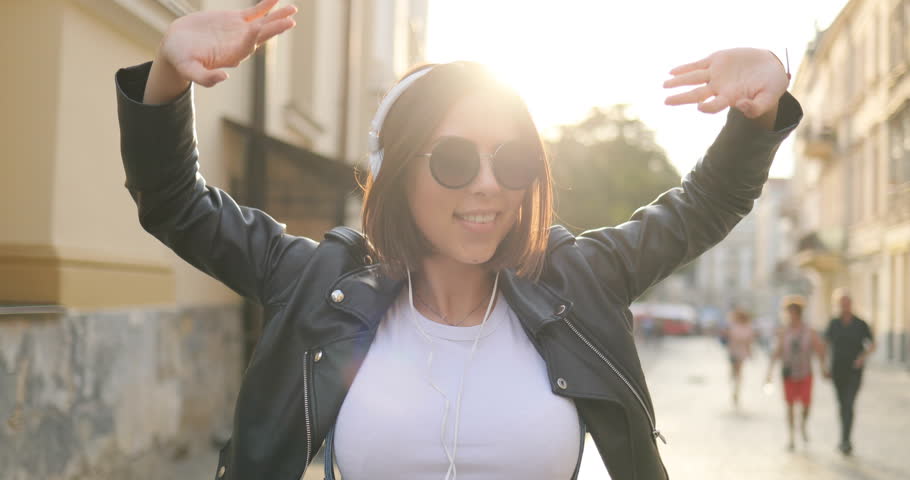 A girl in the headphones is dancing on the street. Happy woman in glasses having fun outdoor.
