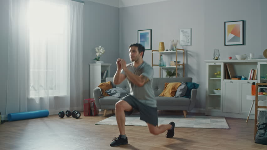 Strong Athletic Fit Man in T-shirt and Shorts is Doing Forward Lunge Exercises at Home in His Spacious and Bright Apartment with Minimalistic Interior. Royalty-Free Stock Footage #1019180710