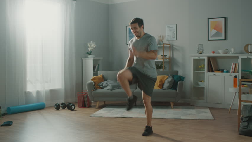 Strong Athletic Fit Man in T-shirt and Shorts is Energetically Jogging in Place at Home in His Spacious and Bright Living Room with Minimalistic Interior. Royalty-Free Stock Footage #1019180734