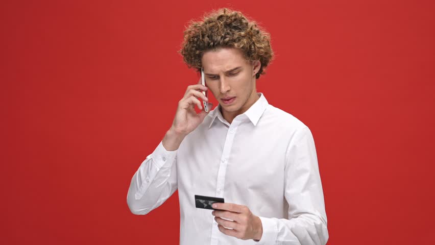 Serious curly man in business shirt talking by smartphone and holding credit card over red background