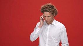 Displeased curly man in business shirt talking by smartphone over red background