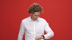 Displeased curly man in business shirt using wristwatch over red background