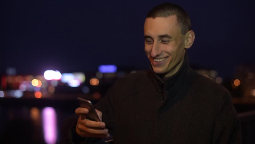 Man smiling while taking selfie or photo of view outdoors at night light. Man sms texting using app on smart phone at night in city. Handsome young student using smartphone smiling happy wearing suit 