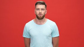 Scared bearded man in t-shirt covering his mouth and looking at the camera over red background
