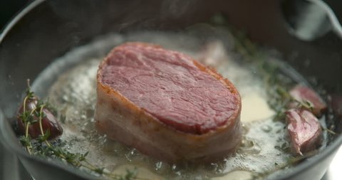 Bacon-wrapped beef tournedos, cooked in butter, garlic and thyme, in a deep frying pan. Close up with shallow depth of field, shot in 4K slow motion with Phantom Flex camera.