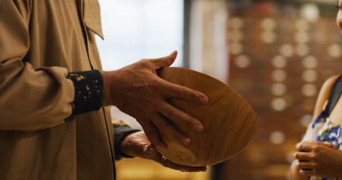 Young Asian woman uses credit card to buy wood bowl from older woodworking artisan man in workshop during the day. Medium to closeup on 4K RED camera.