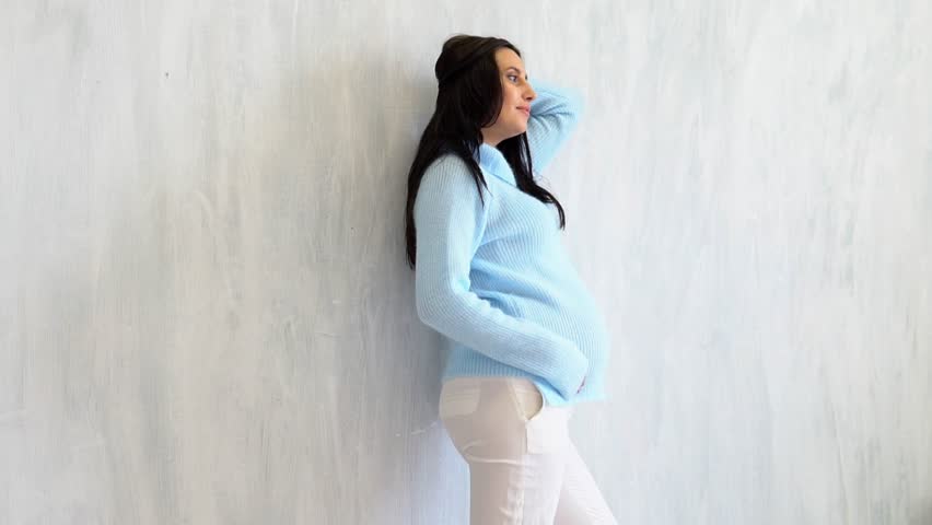 a pregnant woman in a blue sweater stands at white walls
