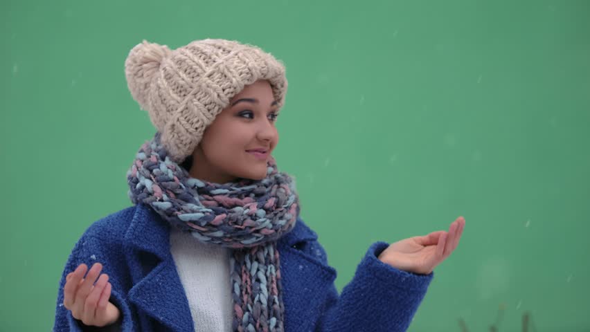 Happy women facing camera having fun during winter holidays outside. Snow falling in slow motion 4K. Young female enjoying winter day outdoors. Woman in cap, coat, scarf standing against green screen