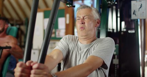 4K Funny gym concept - unfit mature man straining his muscles on weights machine. Slow motion.