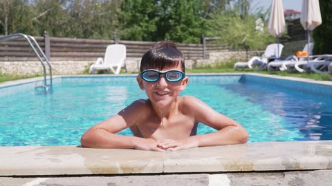 Activity boy in swimming goggles with inflatable orange supports on his arms emerging from the water in pool slow motion. Child clings to the side of pool. Family vacation in hotel