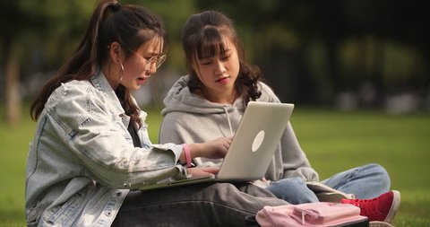 Two happy asian college girl sitting on the lawn using laptop and talking together in campus.
