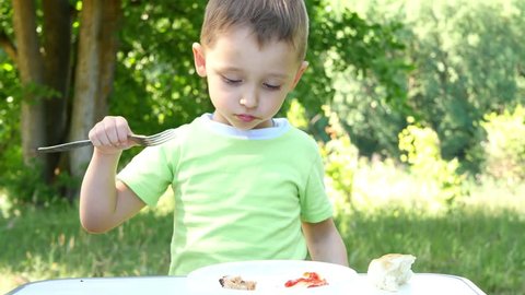 The child eats barbecue, sitting at the table on the background of green forest.