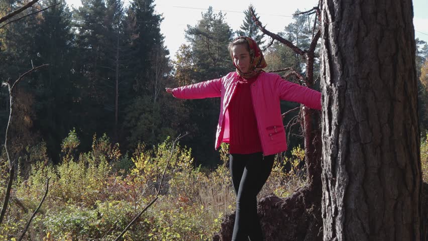 A young, beautiful, rural girl in a pink jacket with a handkerchief on her head is walking on a log, a fallen tree in the forest. She keeps the balance by hand. 