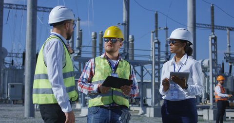 Medium shot of three electrical workers reviewing documents on a tablet during an inspection against the background of a transformer station