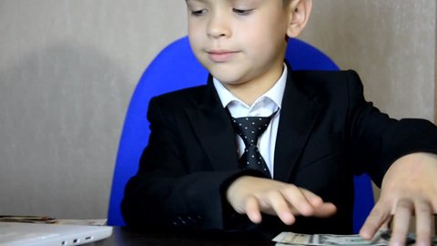 Young businessman using a laptop. Fashion child handsome boy in modern office. sits at a table in an armchair and counts money coins, euro, pounds and dollars.