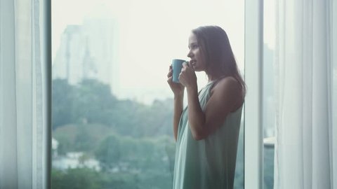 Young beautiful brunette woman opens curtains drinking coffee by the window in her cozy home and looks on the city view. Relaxed morning at home concept. slwo motion. 3840x2160