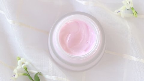 Closeup open moisturizer pink cream in white container on white fabric background with flower, woman use finger applying it by whirl and pat, top view, concept cosmetic