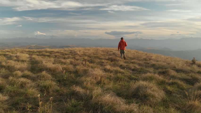 An aerial view of a bearded hipster male with a remote from a drone walks along the edge of a high plateau near a cliff at sunset. The guy walks along the cliff
