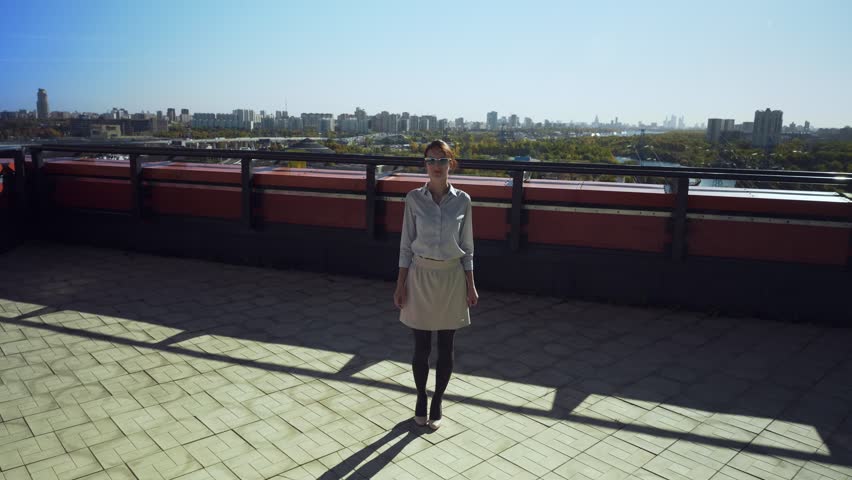 Young business woman in sunglasses with triumph walking on camera on high floor commercial building balcony.