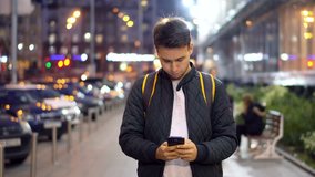 Young Attractive Man Typing Message Using Smartphone in the evening city street with bokeh light effect. 4K Video Clip