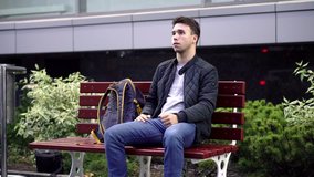 Young man sitting on bench in the street and waiting for something. 4k Video Clip