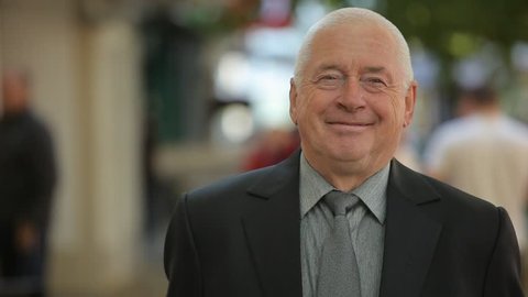  Jolly bokeh portrat of a smiling elderly man in a neat business suit with a grey necktie standing and smiling in a green street in autumn
