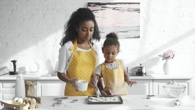 concentrated african american kid in apron brushing dough at tray while her mother standing near in kitchen
