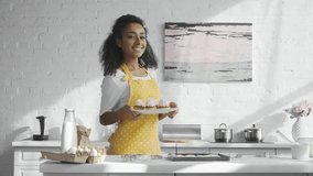 cheerful african american woman in apron holding plate and showing cupcakes in kitchen