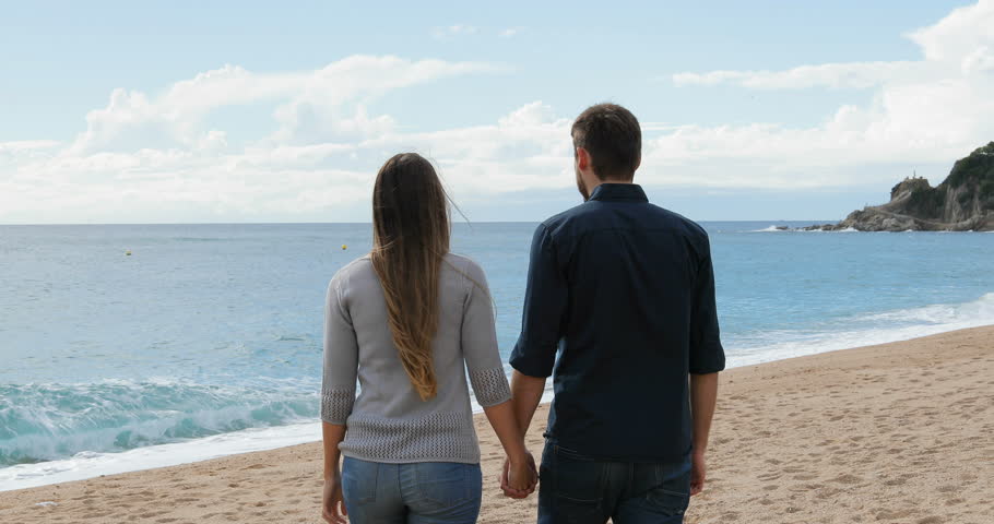 Back view of a happy couple walking holding hands on the beachin a sunny day